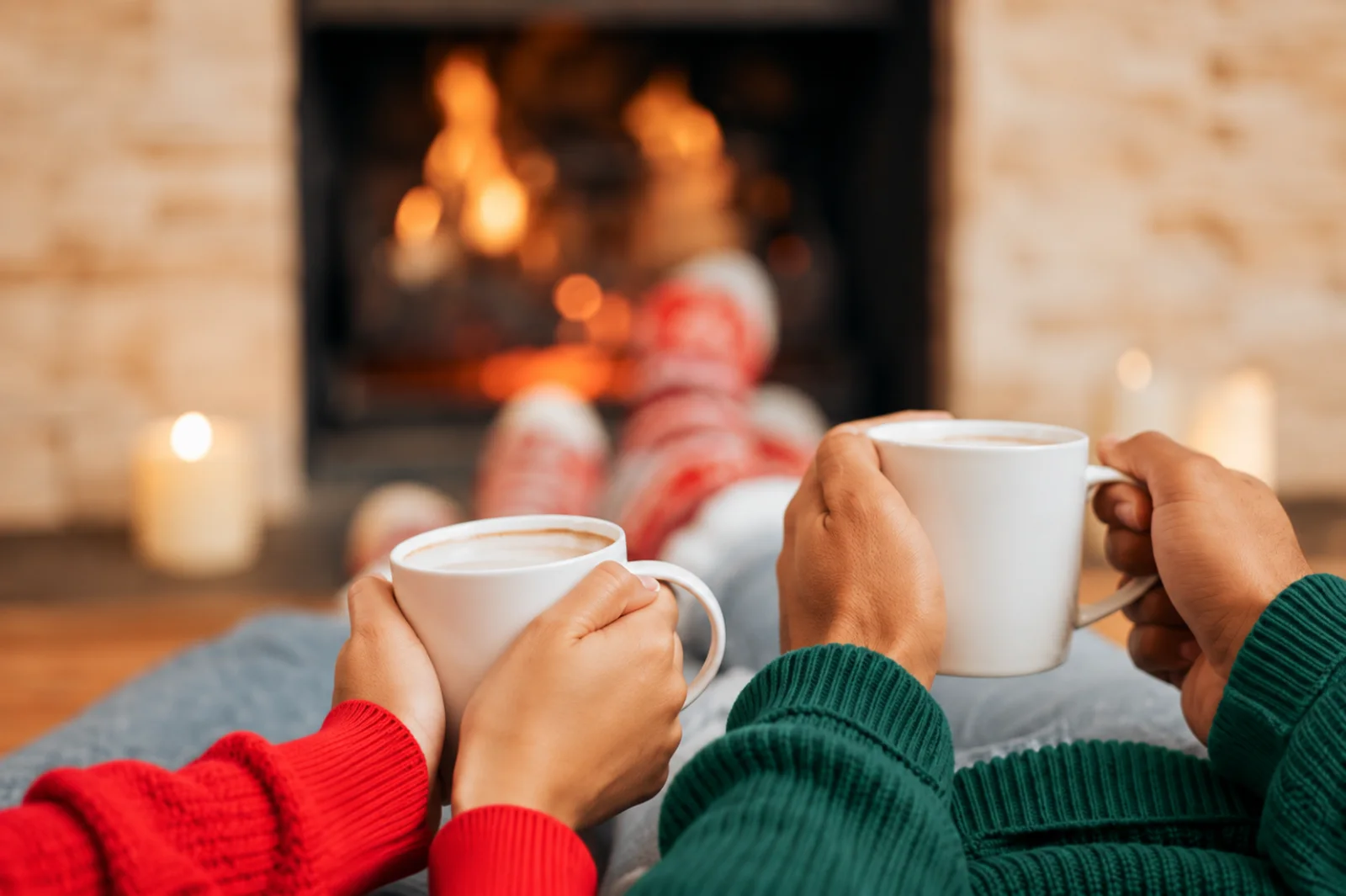 Two people in red and green sweaters holding mugs of cocoa with their feet up in front of a burning fireplace